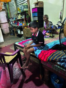 Junge sitzt auf einem Bett, eine ältere Frau ist im Hintergrund