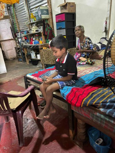 SriLanka_Ditwah_004_web Kind sitzt auf seinem Bett, hinter ihm eine ältere Frau. In dem Raum ist noch alles gestapelt und improvisiert.