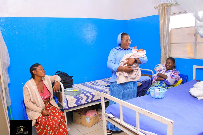 Schwester mit Baby auf dem Arm. Die Mutter und eine Freundin schauen erfreut zu.